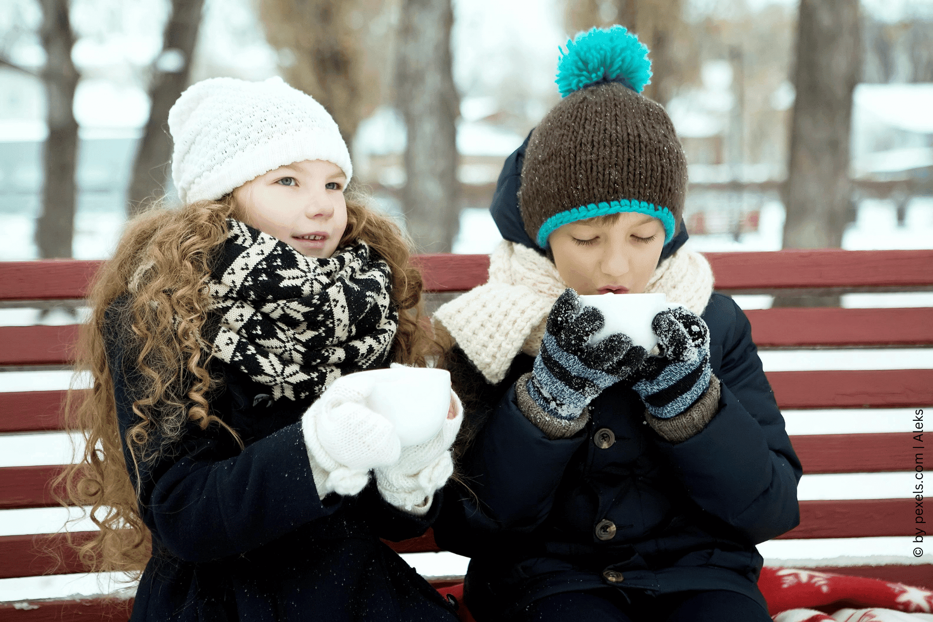 Zwei Kinder in Winterkleidung genießen eine Tasse heiße Schokolade von Tiroler Edle. Sie sitzen auf einer Bank im Schnee.